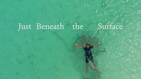 Just Beneath the Surface, Jimmy Thaiday a Torres Strait Islander man lies on his back in clear ocean water, a ghost net is tangled in his body. 