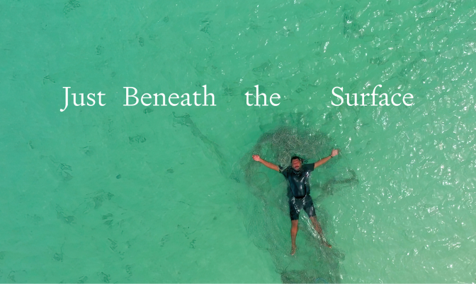 Just Beneath the Surface, Jimmy Thaiday a Torres Strait Islander man lies on his back in clear ocean water, a ghost net is tangled in his body. 
