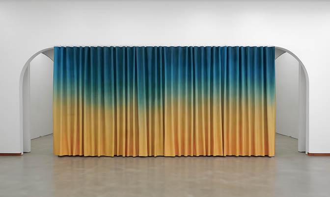 Photo of a vibrantly coloured curtain hanging across arched doorways. The curtain is blue blending into yellow, and the interior walls are white with polished concrete floors. Photo by Ryan Renshaw. Courtesy of the Artist and The Renshaws', Brisbane
